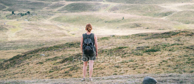 Vista posterior de una excursionista anónima con mochila de pie en la colina en un terreno montañoso y admirando el pintoresco paisaje - foto de stock