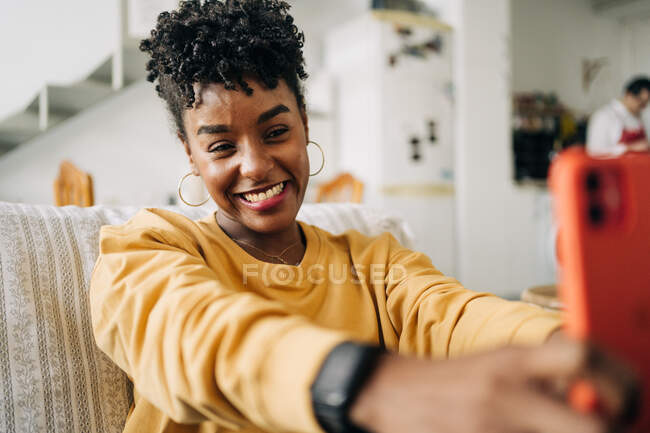 Charmante femme afro-américaine avec des cheveux bouclés prenant autoportrait sur smartphone tout en étant assis sur le canapé à la maison — Photo de stock