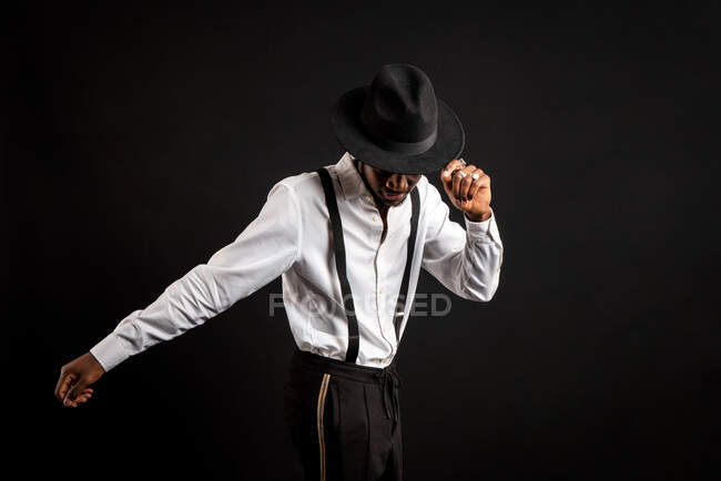 Joven modelo masculino masculino étnico en sombrero y pantalones de pie bailando sobre fondo negro - foto de stock