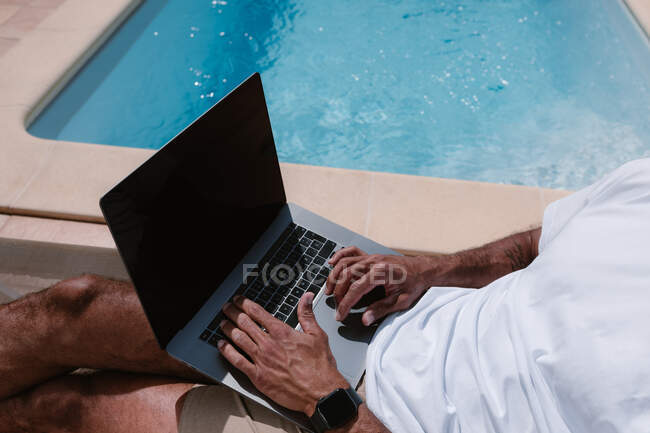 Cortar freelancer masculino irreconhecível deitado na espreguiçadeira à beira da piscina e navegar na Internet no laptop durante o teletrabalho no verão no dia ensolarado — Fotografia de Stock