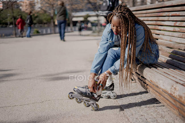 Vista lateral Afro-americana hembra en traje de mezclilla poniéndose patines en día soleado en skate park - foto de stock