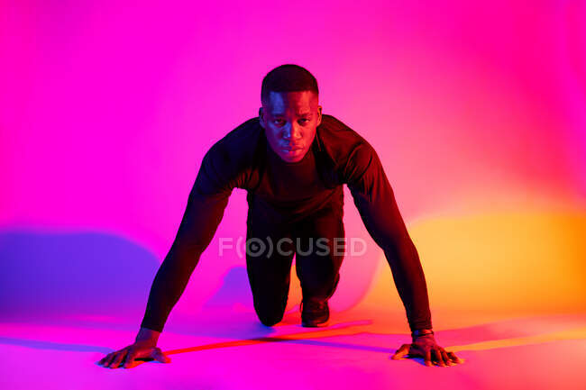 Longitud completa concentrado ajuste afroamericano corredor masculino de pie en posición de inicio agacharse y mirando a la cámara en el fondo colorido en el estudio - foto de stock