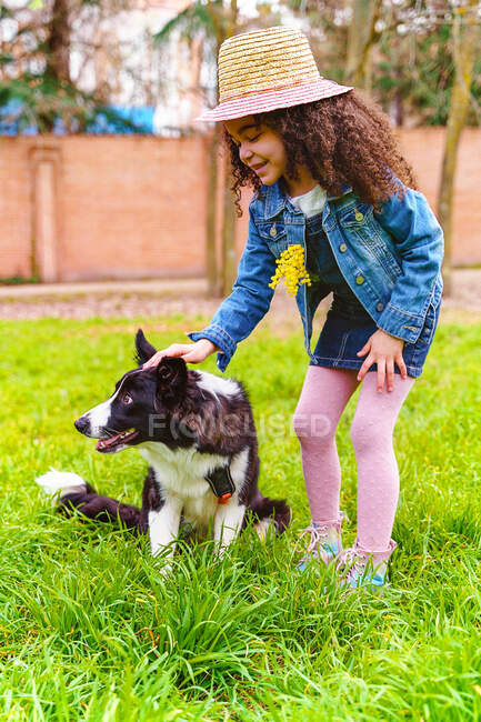 Charming child with curly hair in straw hat stroking adorable purebred dog on lawn in daytime — Stock Photo