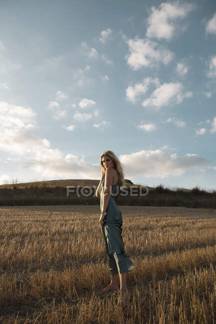 Mujer pacífica en vestido elegante de pie en el campo seco en el área rural y mirando a la cámara - foto de stock