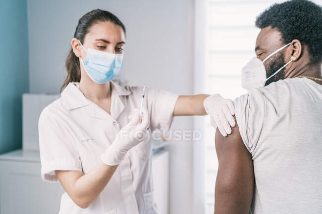 Specialista di medicina femminile in uniforme protettiva, guanti in lattice e maschera facciale che vaccina il paziente afroamericano in clinica durante l'epidemia di coronavirus — Foto stock