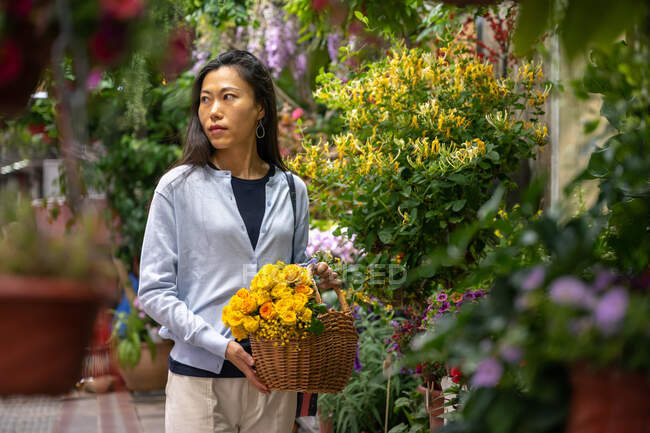 Belle fille asiatique achetant des fleurs dans la boutique de fleurs tout en portant un panier en osier avec des fleurs jaunes. — Photo de stock