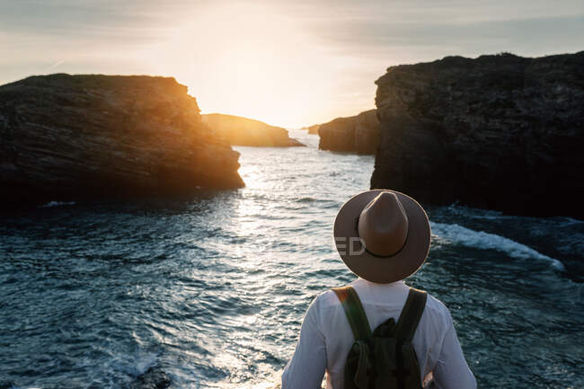 Человек в рюкзаке и шляпе стоит и смотрит на море — стоковое фото