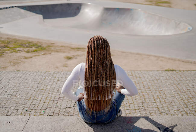 Vista posteriore femmina nera con acconciatura intrecciata e in pattini seduti su rampa in skate park e guardando altrove — Foto stock
