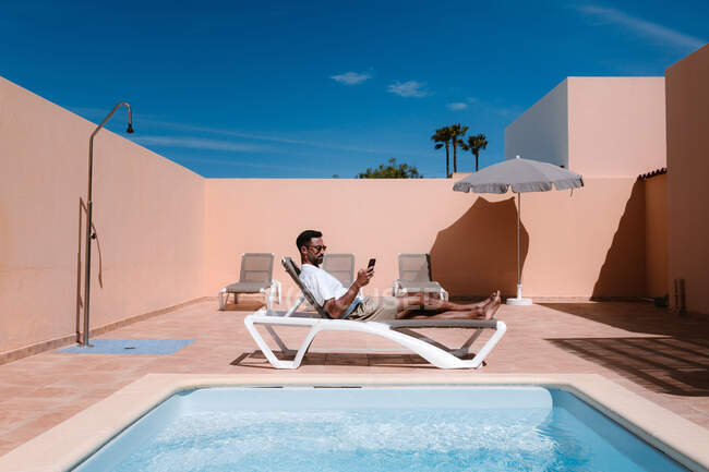Vue latérale d'un homme indépendant allongé sur une chaise longue au bord de la piscine et surfant sur Internet par téléphone portable pendant le télétravail en été par une journée ensoleillée — Photo de stock