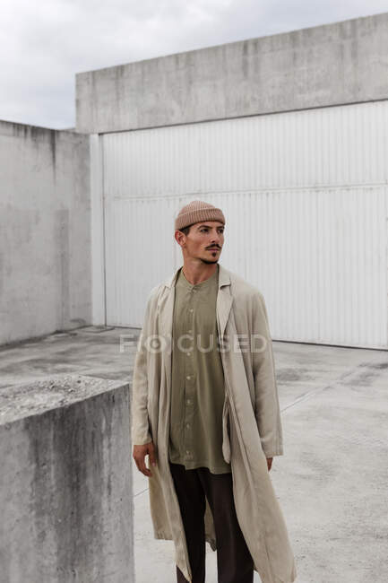 Серьезный мужчина в модном пальто и шляпе стоит в городе и смотрит в сторону — стоковое фото