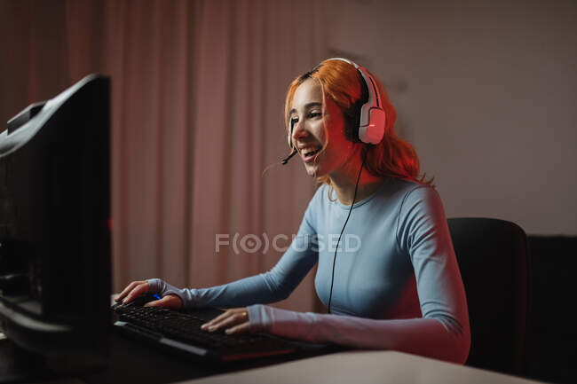 Vista lateral del jugador femenino encantado en auriculares jugando videojuego mientras está sentado en la mesa en casa - foto de stock