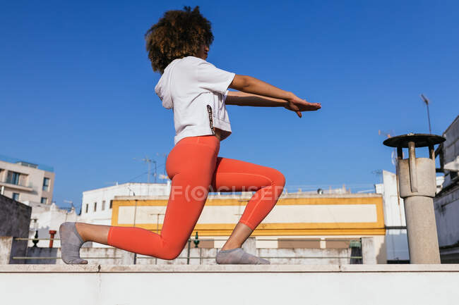Vue latérale à angle bas de la femme afro-américaine calme en vêtements de sport pratiquant le yoga sur le toit le jour ensoleillé — Photo de stock