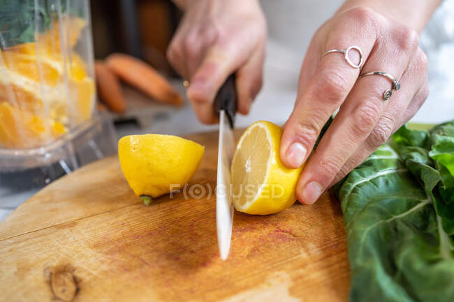 Ritaglio anonimo femminile taglio maturo limone succoso con coltello tra foglie di bietola e ciotola frullatore in cucina — Foto stock