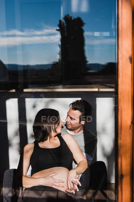 Através da janela da mulher grávida e do homem contente olhando ternamente um para o outro no dia ensolarado em casa — Fotografia de Stock