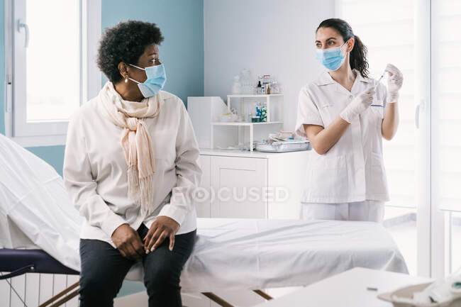 Médecin en gants de latex et bouclier facial remplissant la seringue du flacon avec un vaccin se préparant à vacciner une femme afro-américaine mature méconnaissable en clinique pendant l'épidémie de coronavirus — Photo de stock