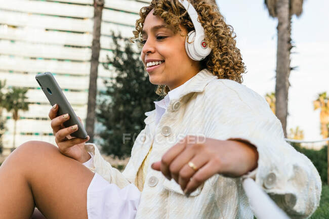 Cheerful African American female browsing mobile phone and listen to music on headphones while sitting on grass in park — Stock Photo