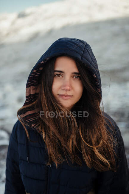 Joven viajero femenino suave en la capucha mirando a la cámara contra las montañas nevadas en invierno en el día soleado - foto de stock