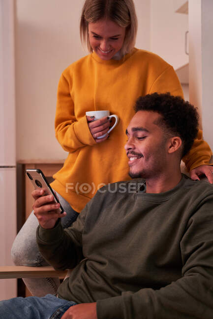 Fröhliches multiethnisches Paar, das mit dem Smartphone surft und Getränke trinkt, während es den Abend zu Hause zusammen genießt — Stockfoto