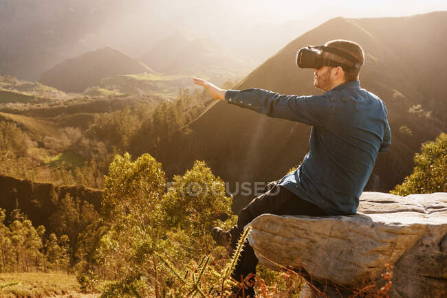 Vista laterale del viaggiatore maschile in occhiali VR che interagisce con la realtà virtuale mentre è seduto sulla collina in un terreno montagnoso al tramonto — Foto stock