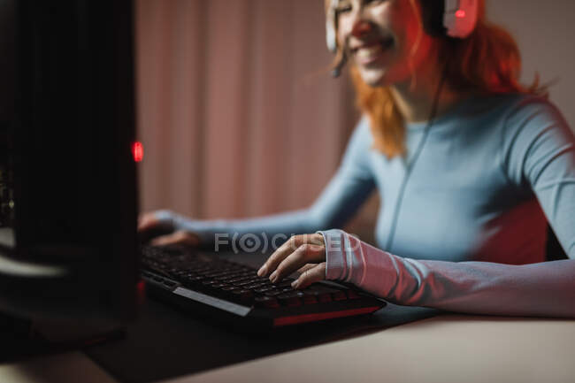 Vista lateral del jugador femenino irreconocible recortado en auriculares jugando videojuego mientras está sentado en la mesa en casa - foto de stock
