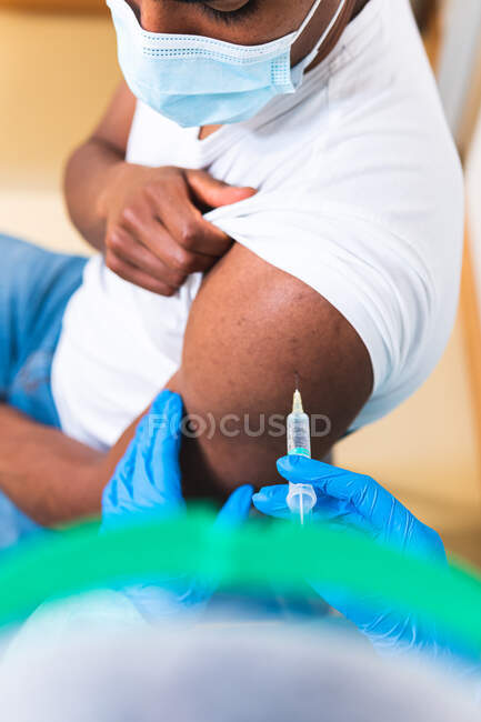 Especialista médica femenina irreconocible en uniforme protector, guantes de látex y mascarilla facial vacunando a un paciente afroamericano en la clínica durante el brote de coronavirus - foto de stock