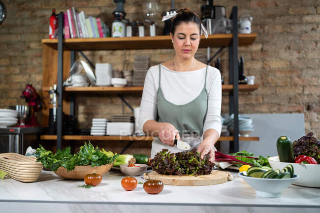 Счастливая женщина режет вкусный овощной салат с листьями салата за столом в лофт стиле дома — стоковое фото