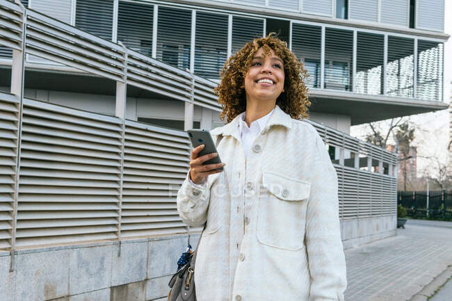 Mujer afroamericana optimista con peinado afro navegando en el teléfono inteligente mientras está de pie contra la pared metálica en el área urbana de la ciudad - foto de stock