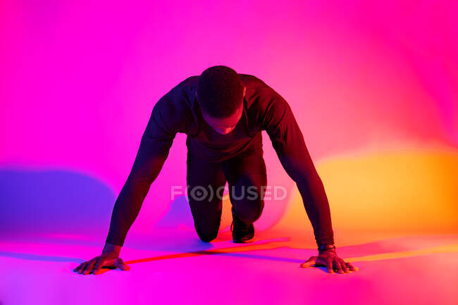 Coupe concentrée pleine longueur coureur mâle afro-américain debout en position de départ accroupi et regardant vers le bas sur fond coloré en studio — Photo de stock