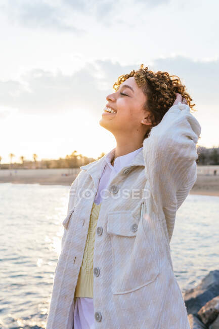 Alto ángulo de alegre hembra afroamericana de pie con los brazos extendidos en las rocas en la orilla del mar y disfrutando de la libertad al atardecer - foto de stock