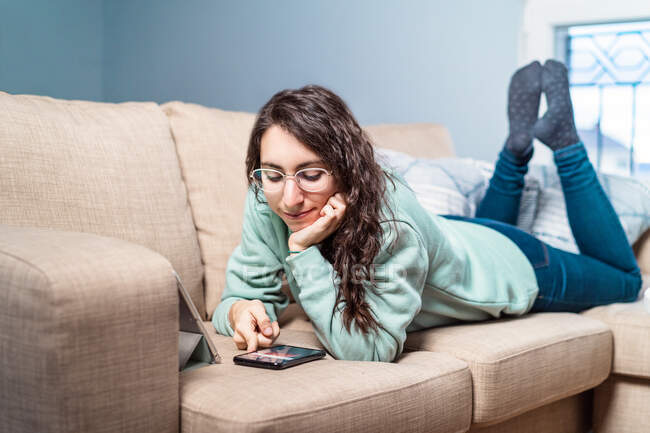 Junge glücklich lächelnde Frau mit türkisfarbenem Sweatshirt und Brille, die sich mit dem Handy auf das Sofa legt — Stockfoto