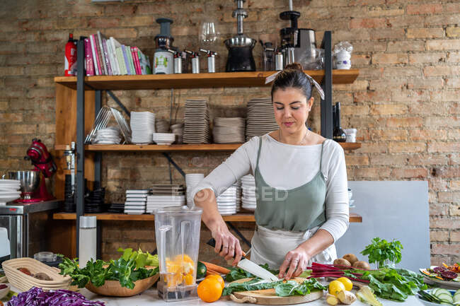 Female cutting fresh chard leaves on chopping board against blender bowl with orange slices in house kitchen — Stock Photo