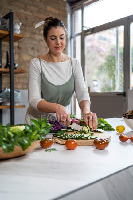 Jovem fêmea cortando abobrinha com faca enquanto prepara o almoço na mesa da cozinha em casa — Fotografia de Stock