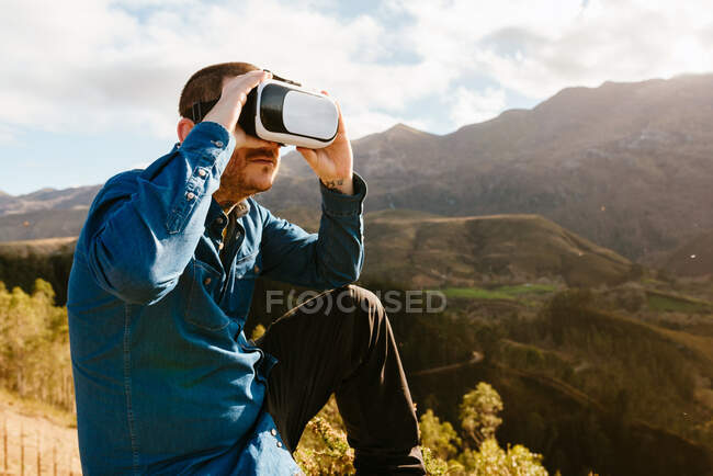 Curioso viajero masculino sentado en la colina y experimentando la realidad virtual en gafas en las montañas en un día soleado - foto de stock