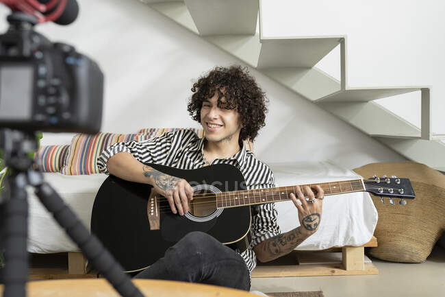 Junger tätowierter männlicher Gitarrist spielt Akustikgitarre, während er Video auf Fotokamera im Hauszimmer aufnimmt — Stockfoto