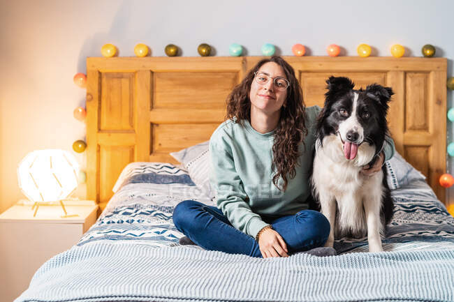 Young woman in glasses sitting on bed hugging border collie dog looking at camera — Stock Photo