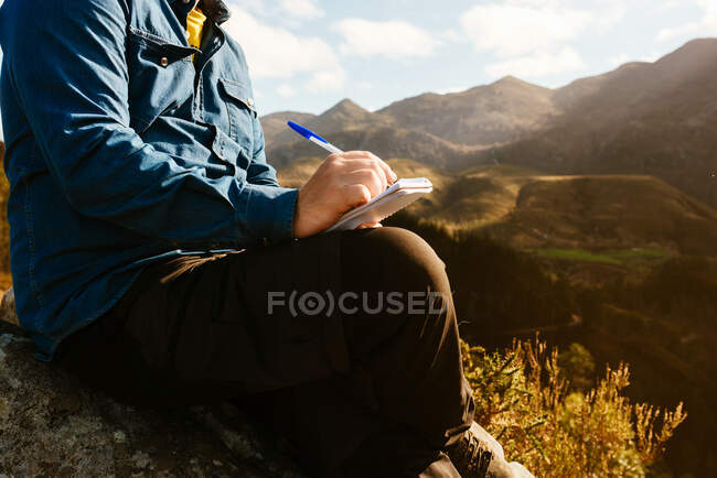 Side view of crop unrecognizable male explorer sitting on hill in mountains and writing in notepad during vacation — Stock Photo