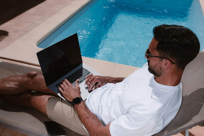 Мужчина-фрилансер лежит на шезлонге у бассейна и занимается серфингом в Интернете на ноутбуке во время телеработы летом в солнечный день — стоковое фото