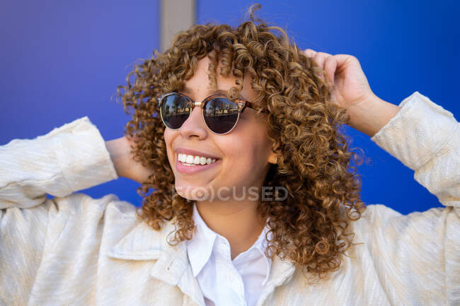 Deliziosa donna afroamericana con acconciatura riccia che indossa occhiali da sole in piedi su sfondo blu in studio — Foto stock