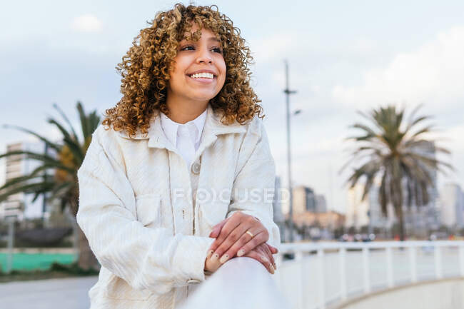 Side view of carefree African American female with curly hair leaning on fence and looking away in city in evening — Stock Photo