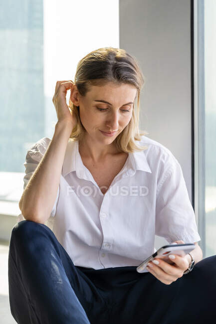 Одинокая неэмоциональная молодая женщина сидит в пустом офисе с большим окном просматривает на мобильном телефоне — стоковое фото