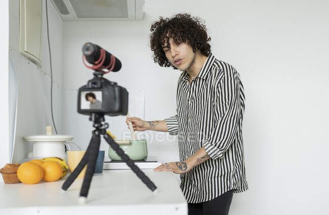 Jeune homme en chemise rayée parlant contre appareil photo sur trépied pendant le processus de cuisson dans la cuisine — Photo de stock