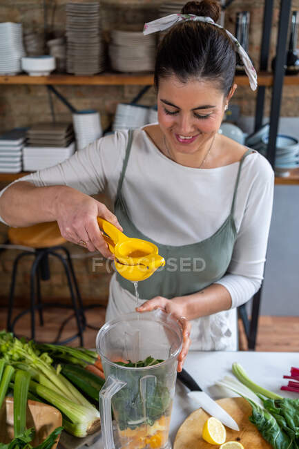 Conteúdo feminino espremendo suco de limão em folhas de acelga em tigela de liquidificador enquanto prepara bebida saudável na cozinha da casa — Fotografia de Stock