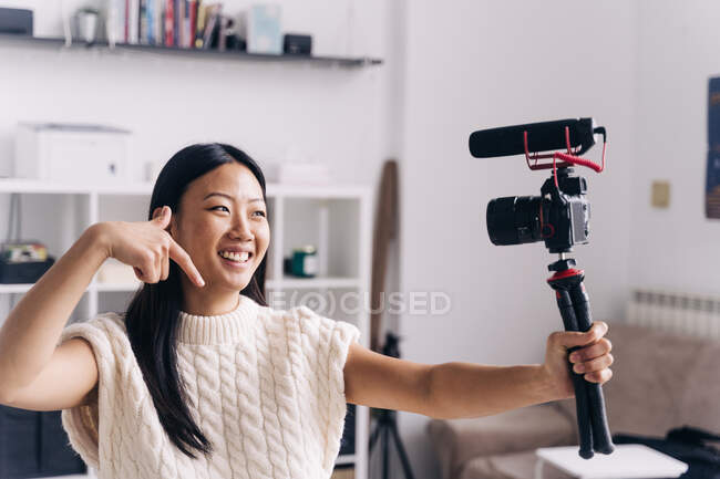 Smiling ethnic female vlogger recording video on photo camera while making hand gestures standing in living room — Stock Photo