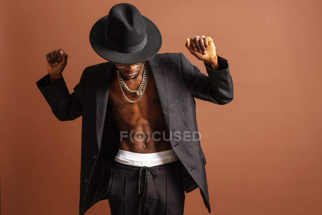 Junger maskuliner unrasierter afroamerikanischer Mann mit nacktem Bauch in Jacke, der auf braunem Hintergrund nach unten schaut — Stockfoto