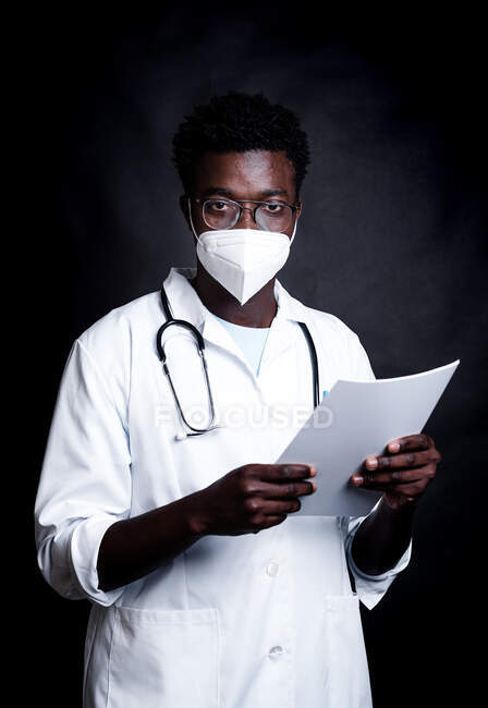 Афроамериканський лікар у захисній масці та біла уніформа роблять нотатки на дошці, дивлячись на камеру на чорному фоні. — стокове фото