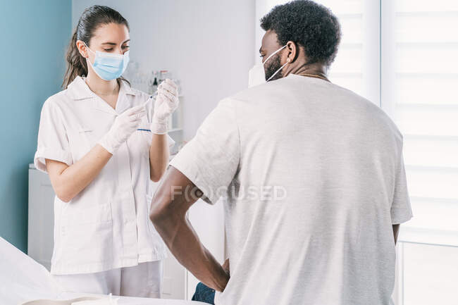 Médecin en gants de latex et bouclier facial remplissant la seringue du flacon avec un vaccin se préparant à vacciner un patient afro-américain méconnaissable en clinique pendant l'épidémie de coronavirus — Photo de stock