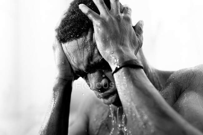 Noir et blanc de jeune homme noir sans émotion prenant une douche dans la salle de bain lumineuse avec les yeux fermés et de l'eau sur le visage — Photo de stock