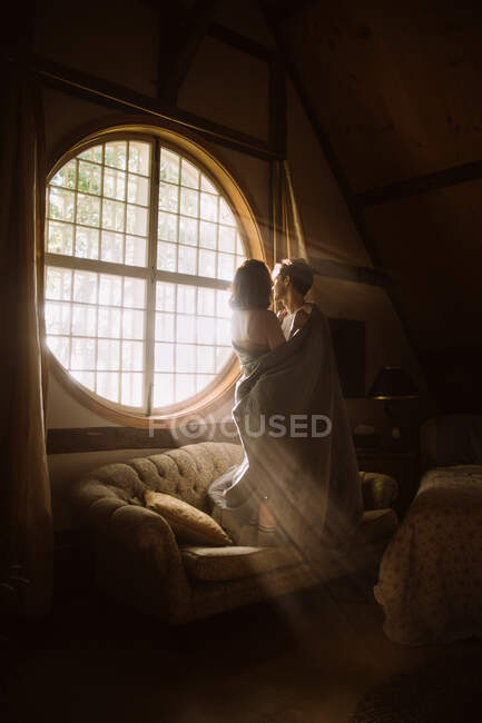 Homem anônimo abraçando namorada com têxteis no sofá enquanto olha para fora em forma de janela em luz solar — Fotografia de Stock