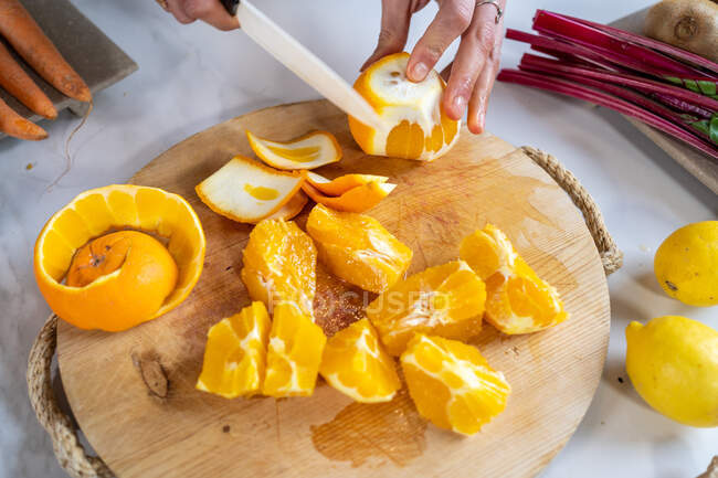 Неузнаваемая женщина очищает спелые апельсины ножом на деревянной доске за кухонным столом — стоковое фото
