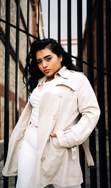 Kleine Plus-Size-Frau im schicken Mantel steht mit den Händen in den Taschen am Zaun in der Stadt und blickt in die Kamera — Stockfoto